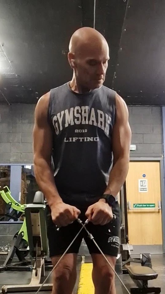 Jason Isaacs at a cable machine in the gym, black vest, both handles pulled to his hips